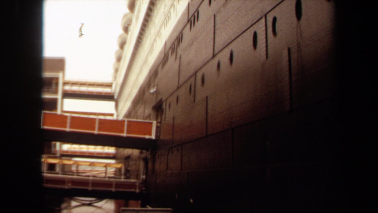 1971 Long Beach home-movie frame looking up at a large dark ship hull with rows of portholes and a red-and-white connector walkway.
