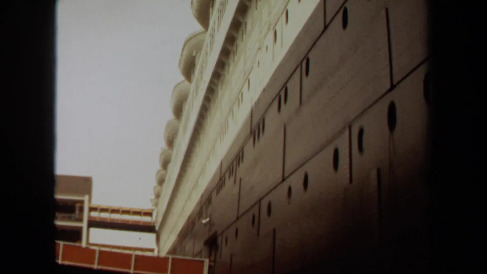 1971 Long Beach home-movie still looking upward along the side of a large ship-like structure with portholes and a partial walkway below.