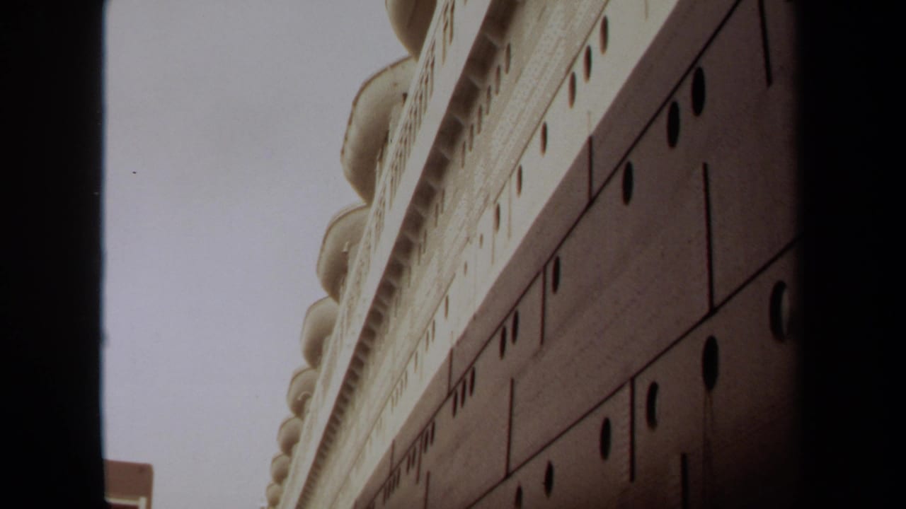1971 Long Beach home-movie frame looking up along a large docked ship, emphasizing its dark hull, portholes, and white superstructure.
