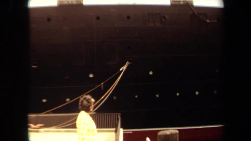 1971 Long Beach home-movie frame with a woman in a yellow patterned dress near the pier railing below the ship's hull and mooring gear.