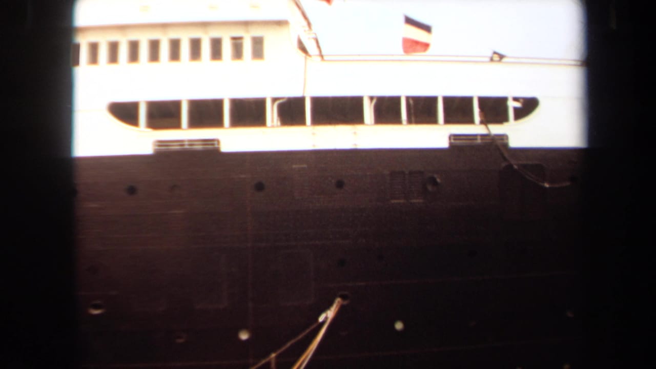 Large ship side with dark hull, white upper deck, windows, and flags above.