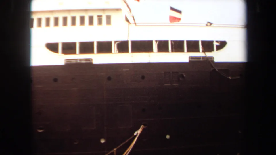 1971 Long Beach home-movie frame showing a docked ocean liner’s dark hull, white superstructure, long window band, and flags along the top deck.