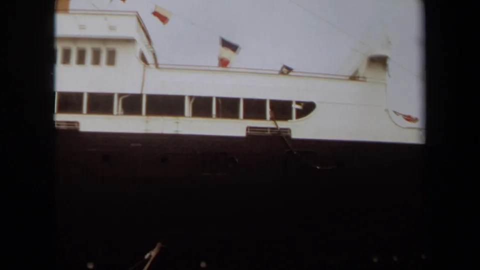 1971 Long Beach home-movie view of the ship’s side, showing the white superstructure, long window strip, dark hull, and flags in the wind.