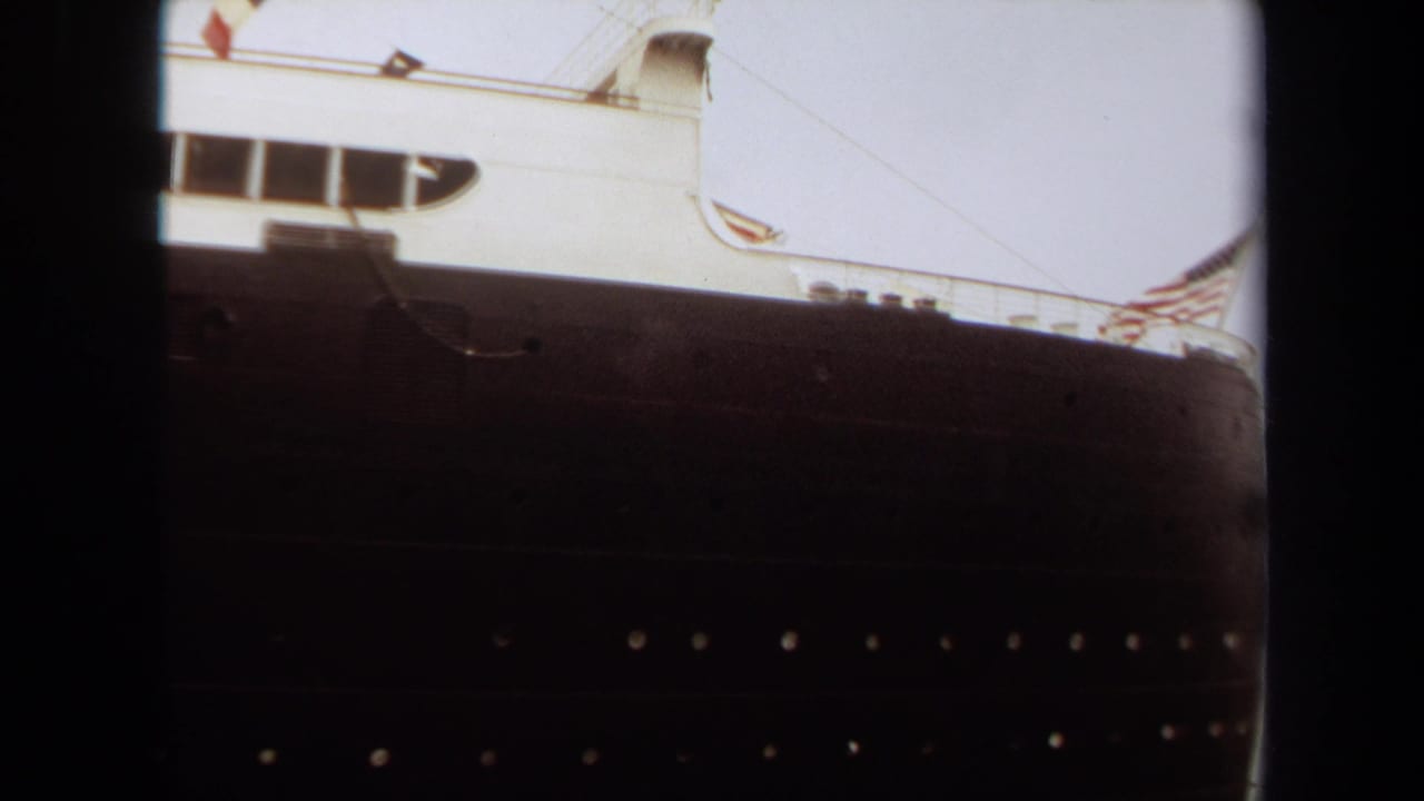1971 8mm view of the Queen Mary docked at Long Beach, showing the dark hull, white superstructure, portholes, and film gate edges.