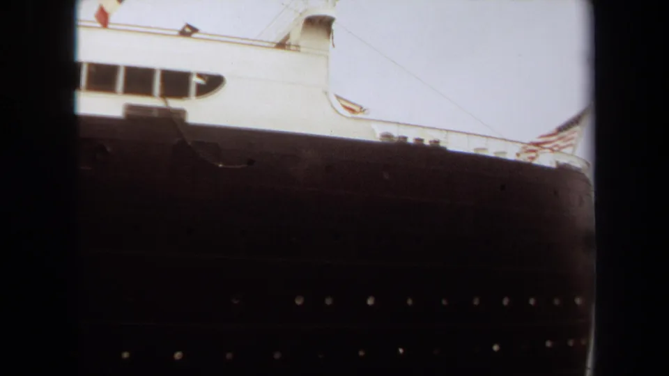 1971 8mm view of the Queen Mary docked at Long Beach, showing the dark hull, white superstructure, portholes, and film gate edges.