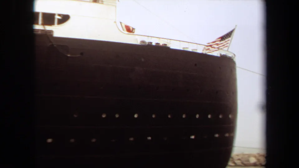 The Queen Mary's bow area appears in 1971 Long Beach footage, with a dark hull, white deck structure, rows of portholes, and an American flag.
