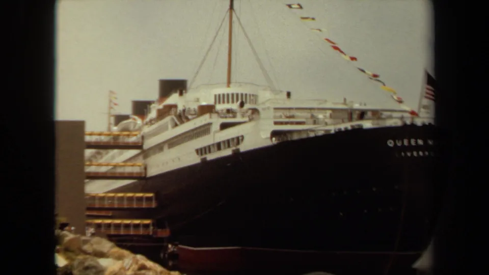 Wider 1971 Long Beach home-movie frame showing the Queen Mary docked, with signal flags, white decks, dark bow, rocks, and harbor edge.
