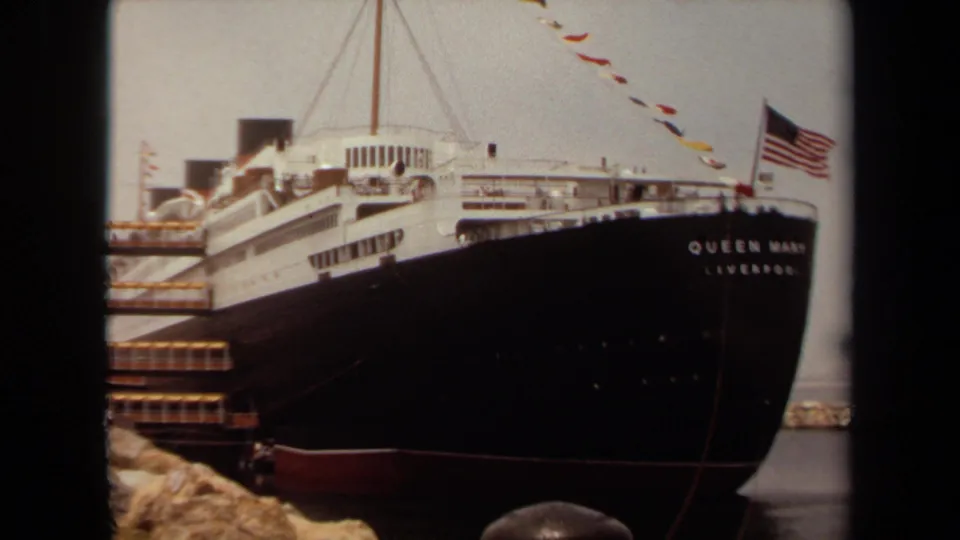 1971 8mm frame from Long Beach showing the Queen Mary name on the bow, American flag, signal flags, dockside rocks, and dark hull.