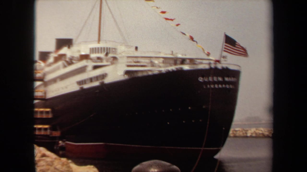 1971 Long Beach home-movie frame showing the Queen Mary bow, American flag, bunting, harbor rocks, and water.