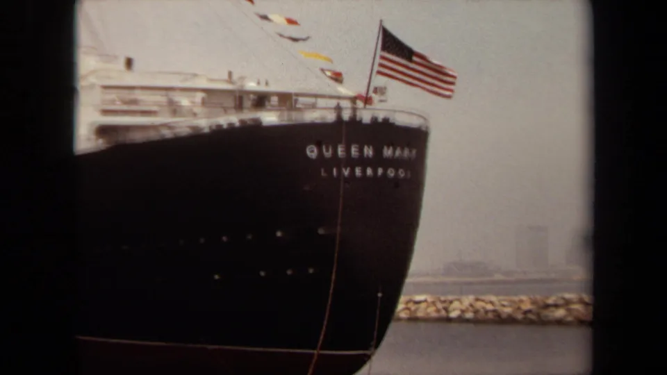 1971 Long Beach 8mm view of the Queen Mary's bow, visible ship lettering, American flag, breakwater, and distant skyline haze.