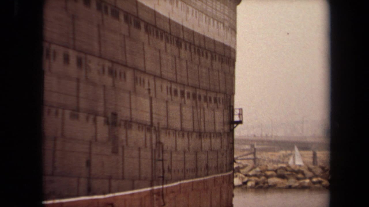 Large ship hull beside harbor water with a distant sailboat near a rocky breakwater.