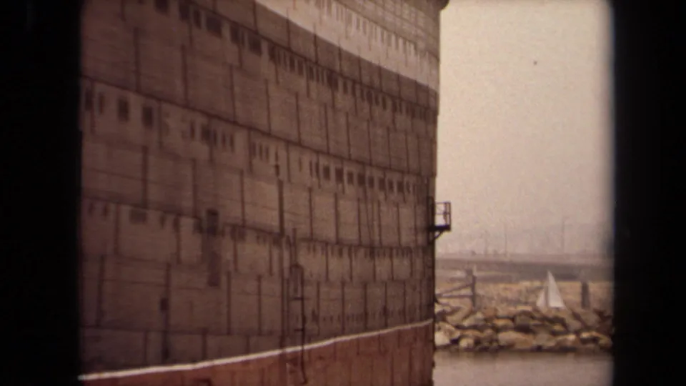 1971 Long Beach home-movie frame showing the dark hull of a large ocean liner with harbor water, breakwater rocks, and a small sailboat beyond.