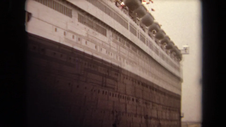 1971 Long Beach 8mm frame looking upward along the side of a large docked liner, with portholes, lifeboat shapes, and deck structures visible.