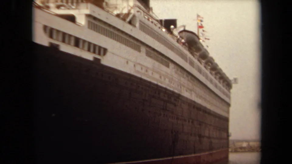 1971 Long Beach frame showing the liner's side from a low angle, with dark hull, white upper decks, flags or deck details, and distant breakwater.