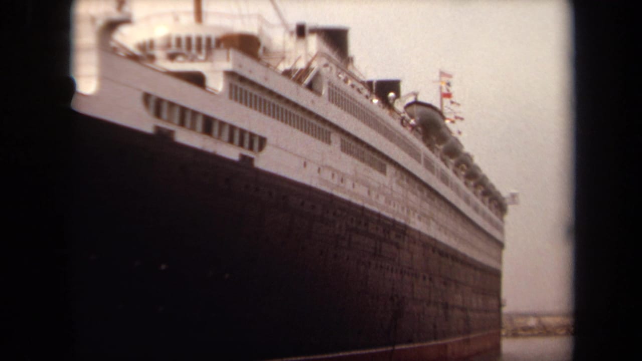 1971 Long Beach 8mm frame showing a massive docked ship from below, with a dark hull, white superstructure, windows, and faint flags.