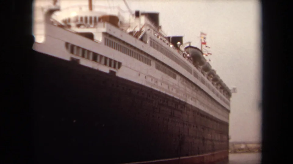 1971 Long Beach 8mm frame showing a massive docked ship from below, with a dark hull, white superstructure, windows, and faint flags.