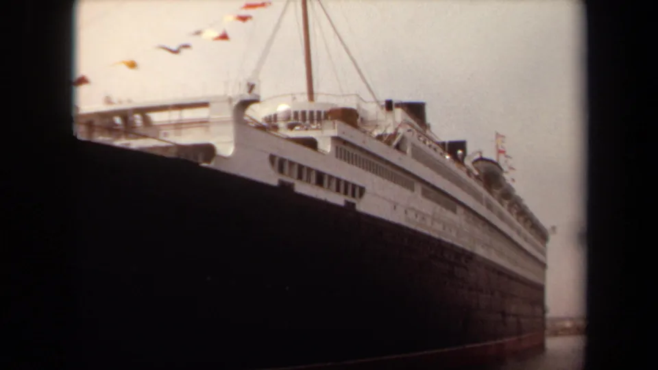 1971 Long Beach 8mm still showing the ocean liner's long side, white decks, dark hull, rigging, and colorful flags near the mast.