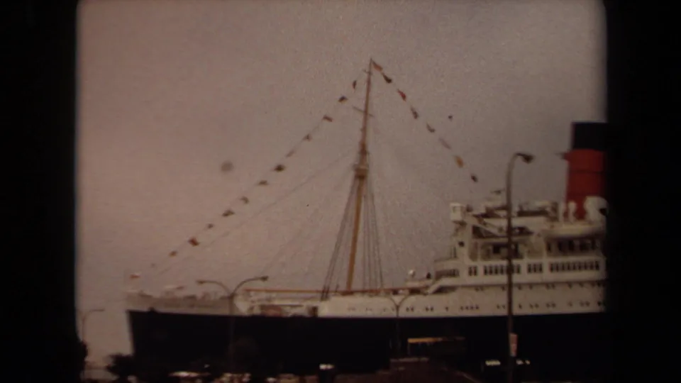 1971 Long Beach frame with a wider harbor view of the ship's forward section, mast rigging, signal flags, and a red-and-black funnel.