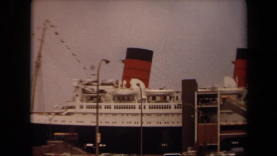 The camera has panned along the docked passenger liner, showing the funnel, rows of deck windows, mast rigging, and gangway area at Long Beach.