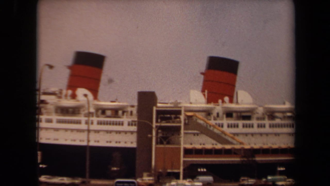 Large white ocean liner docked beside a waterfront structure with red-and-black funnels visible.