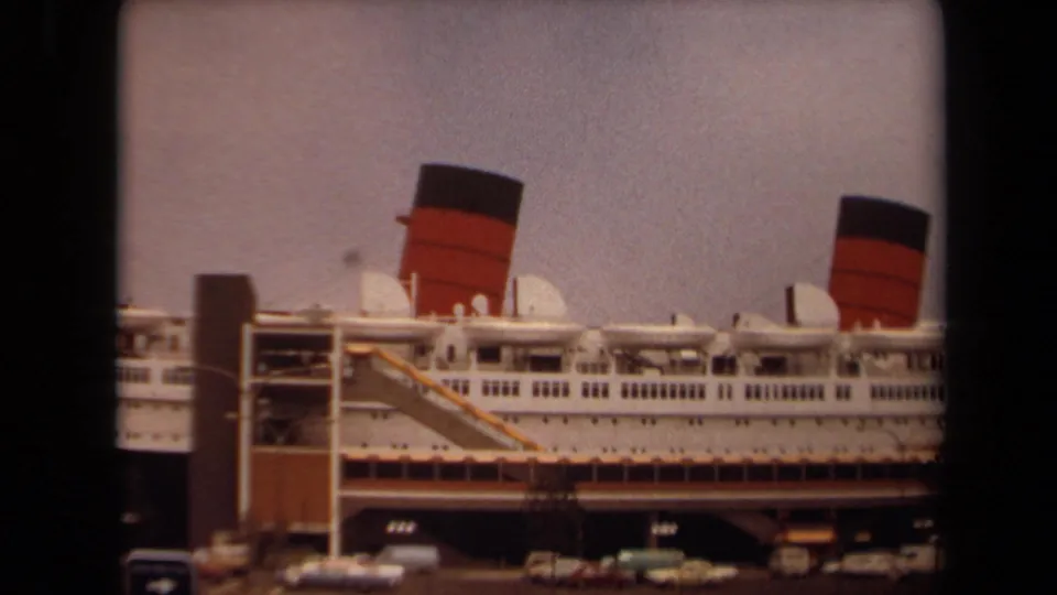 1971 Long Beach view of the liner's side, showing funnels, upper deck details, a dock access structure, and cars below.