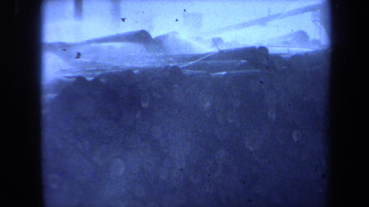 1971 Maine home-movie frame showing stacked wooden logs across a hazy waterfront with tall mast-like poles in the background.
