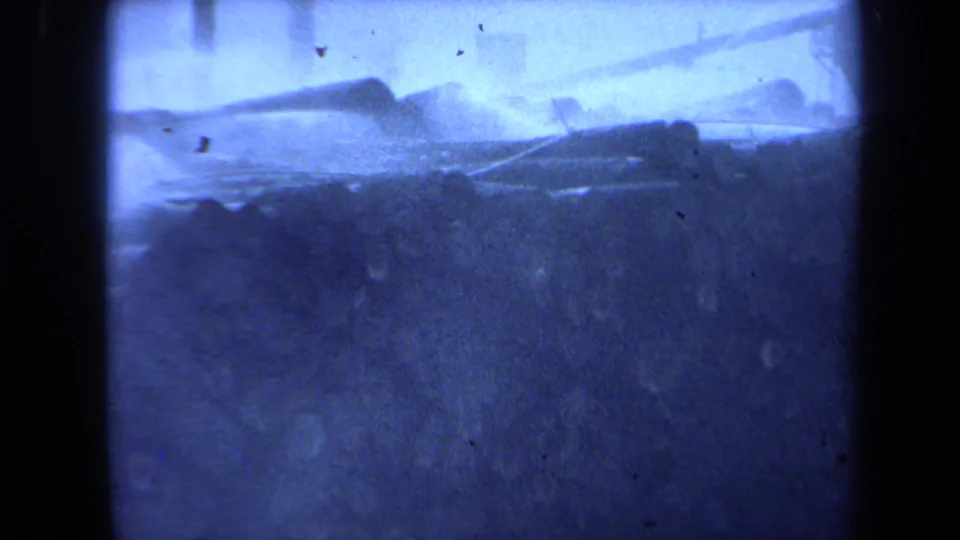 1971 Maine home-movie frame showing stacked wooden logs across a hazy waterfront with tall mast-like poles in the background.