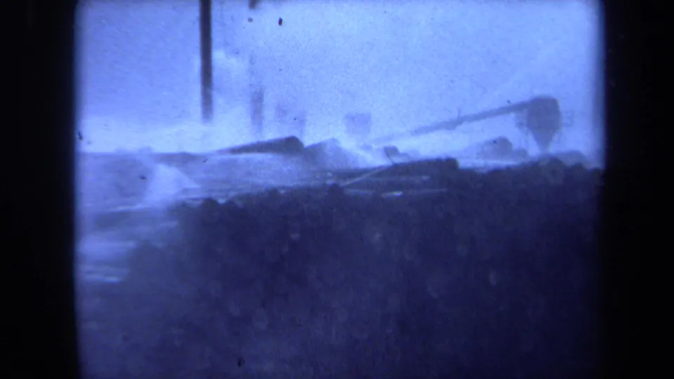 Blue-tinted 8mm view of stacked logs along a foggy Maine waterfront, with tall mast-like forms rising behind them.
