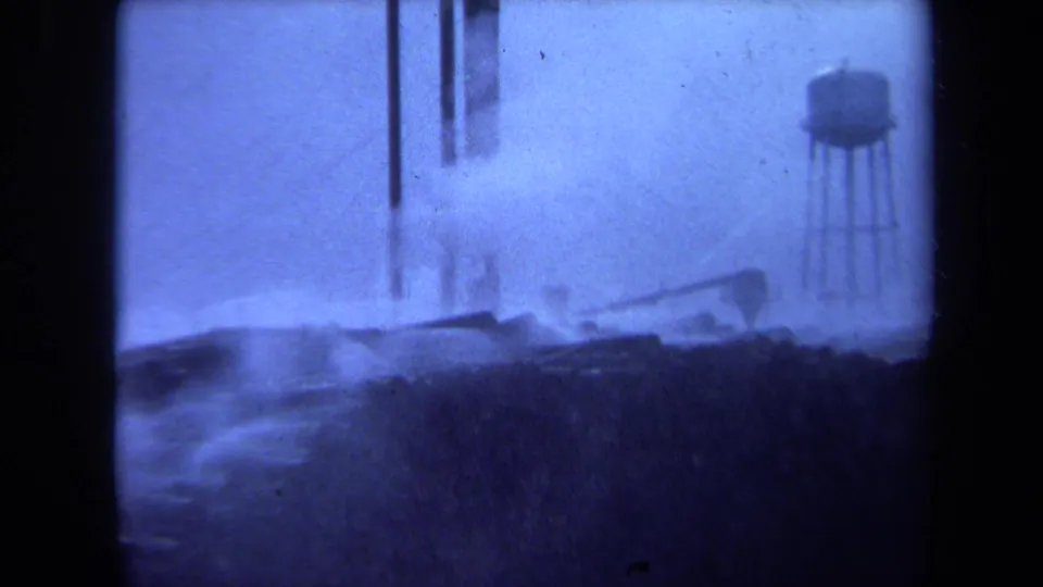 1971 Maine frame with stacked waterfront logs, tall vertical poles, and a distant water tower visible through haze.