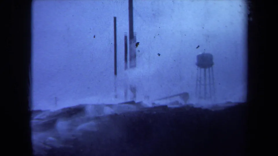 Final 1971 Maine home-movie frame showing logs along a foggy waterfront with mast-like poles and a small water tower in the distance.