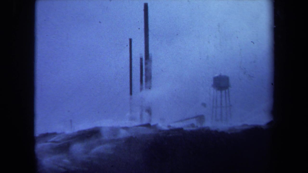 1971 Maine 8mm frame showing a hazy industrial landscape with a water tower, vertical stacks, low structures, and heavy blue film cast.