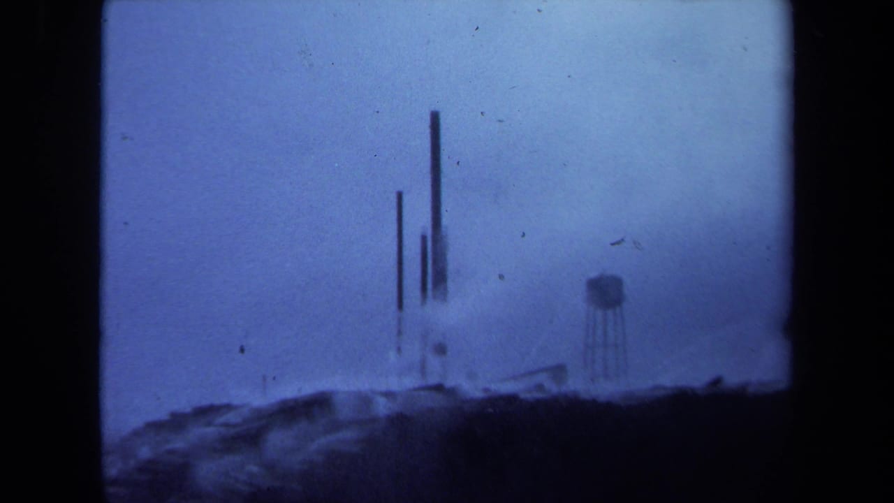 Soft blue view of distant stacks, water tower, low industrial shapes, haze, and dark foreground.