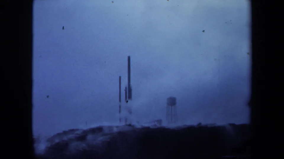 1971 Maine 8mm frame with a hazy industrial horizon, water tower at right, vertical stacks near center, and visible film grain and specks.