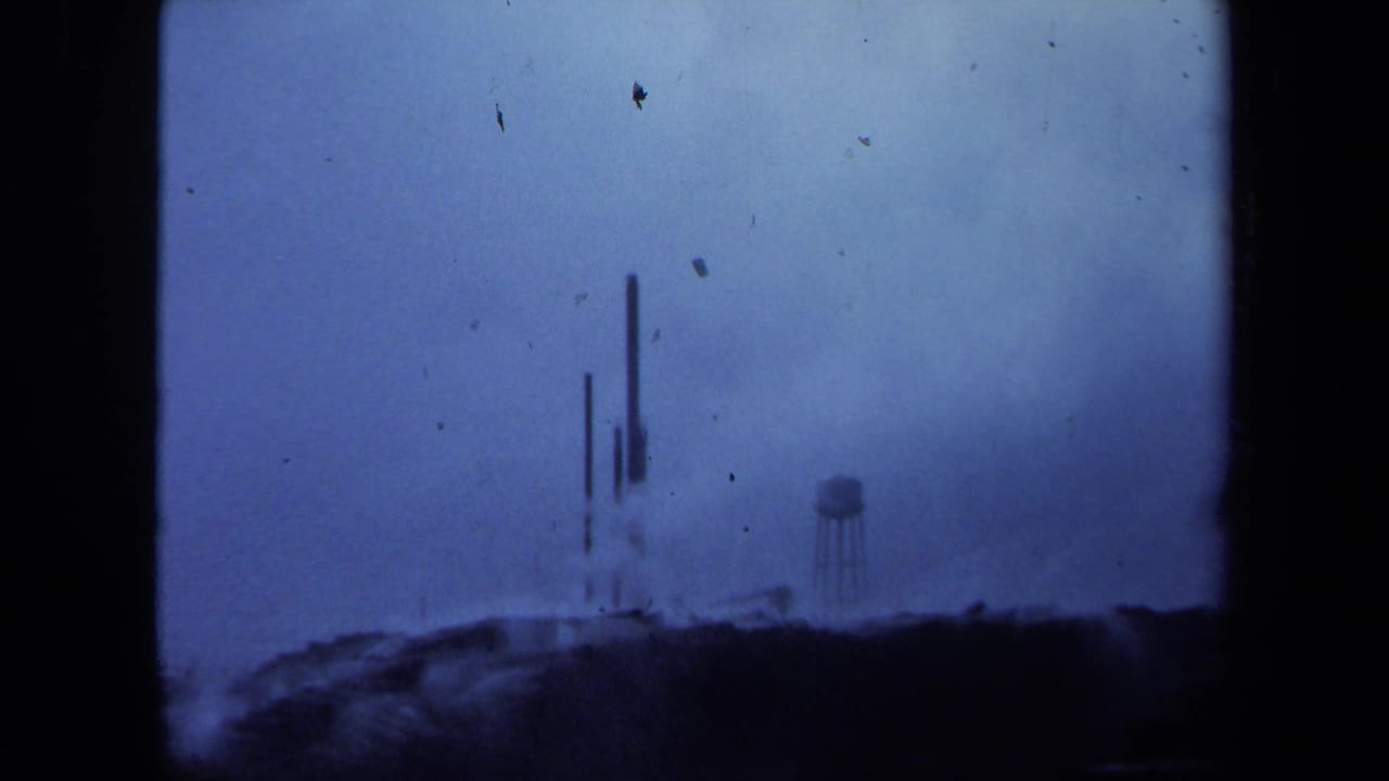 1971 Maine home-movie frame showing a distant industrial landscape with vertical stacks, a water tower, haze, and blue film color shift.