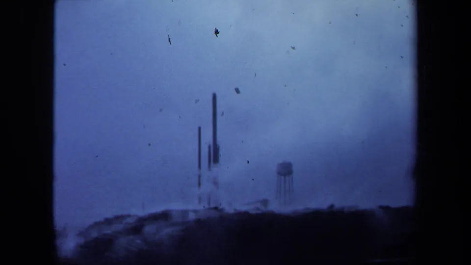1971 Maine home-movie frame showing a distant industrial landscape with vertical stacks, a water tower, haze, and blue film color shift.