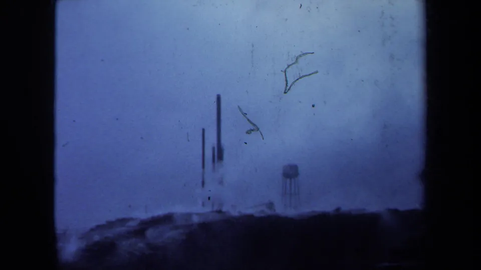 Blue-shifted 1971 Maine frame with vertical industrial stacks, a small water tower, low horizon, dust, and scratches across the film.