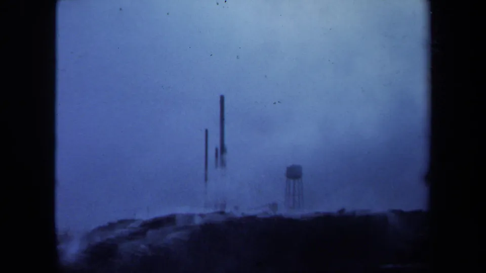 Final 1971 Maine frame holds on the distant industrial site, with stacks, a water tower, haze, dark ground, and soft blue 8mm exposure.