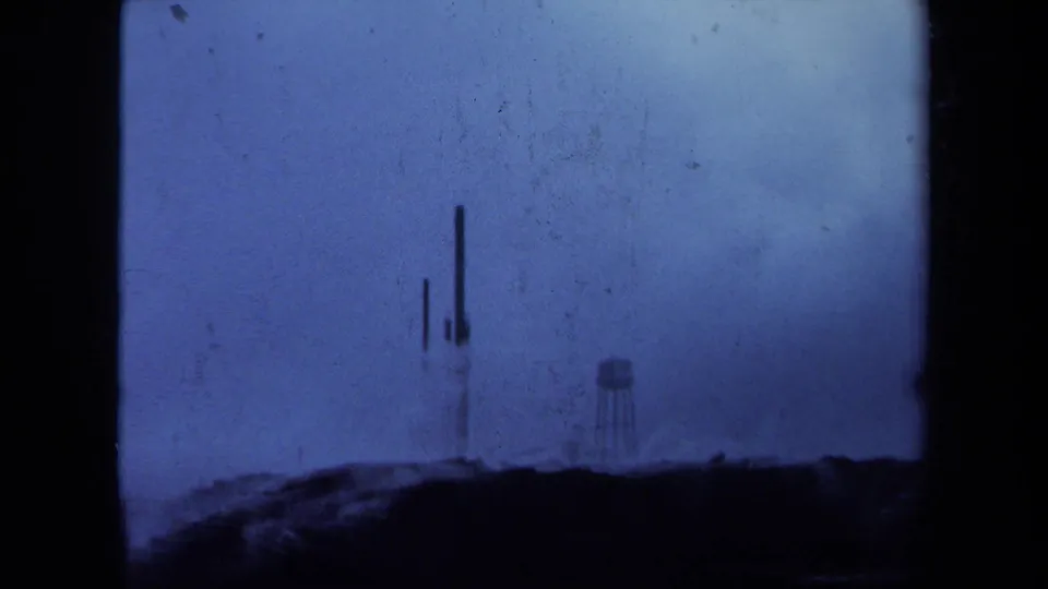 1971 Maine 8mm frame with soft silhouettes of tall metal structures and a water tower against a cloudy sky.