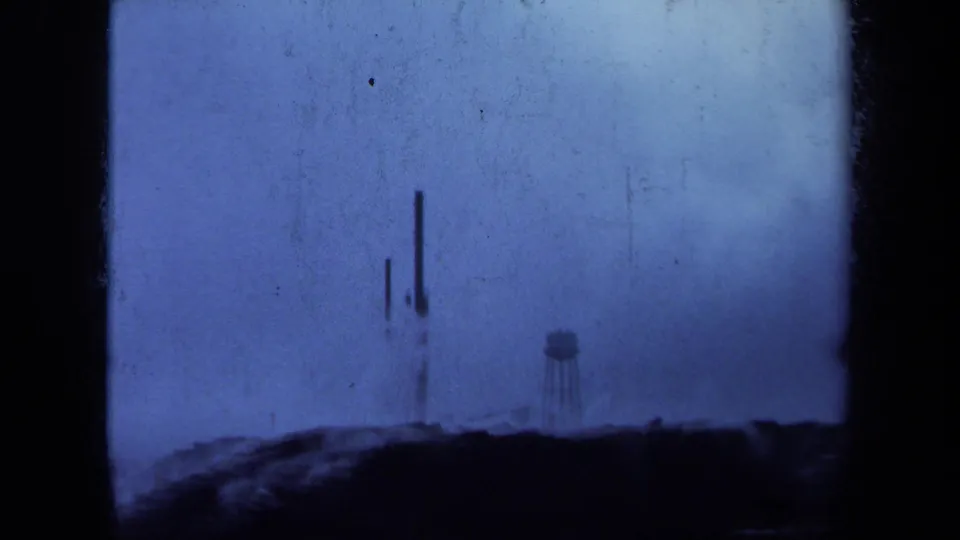 Scratched 1971 Maine 8mm view of tall narrow metal structures and a water tower beyond a dark foreground ridge.