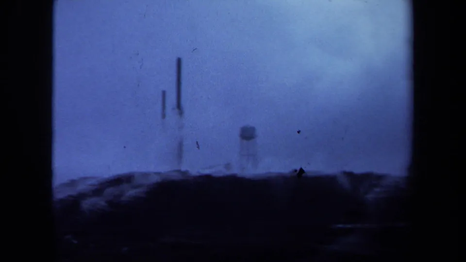 Blurred 1971 Maine 8mm frame of distant towers and a water tower above a dark hillside under pale cloud cover.