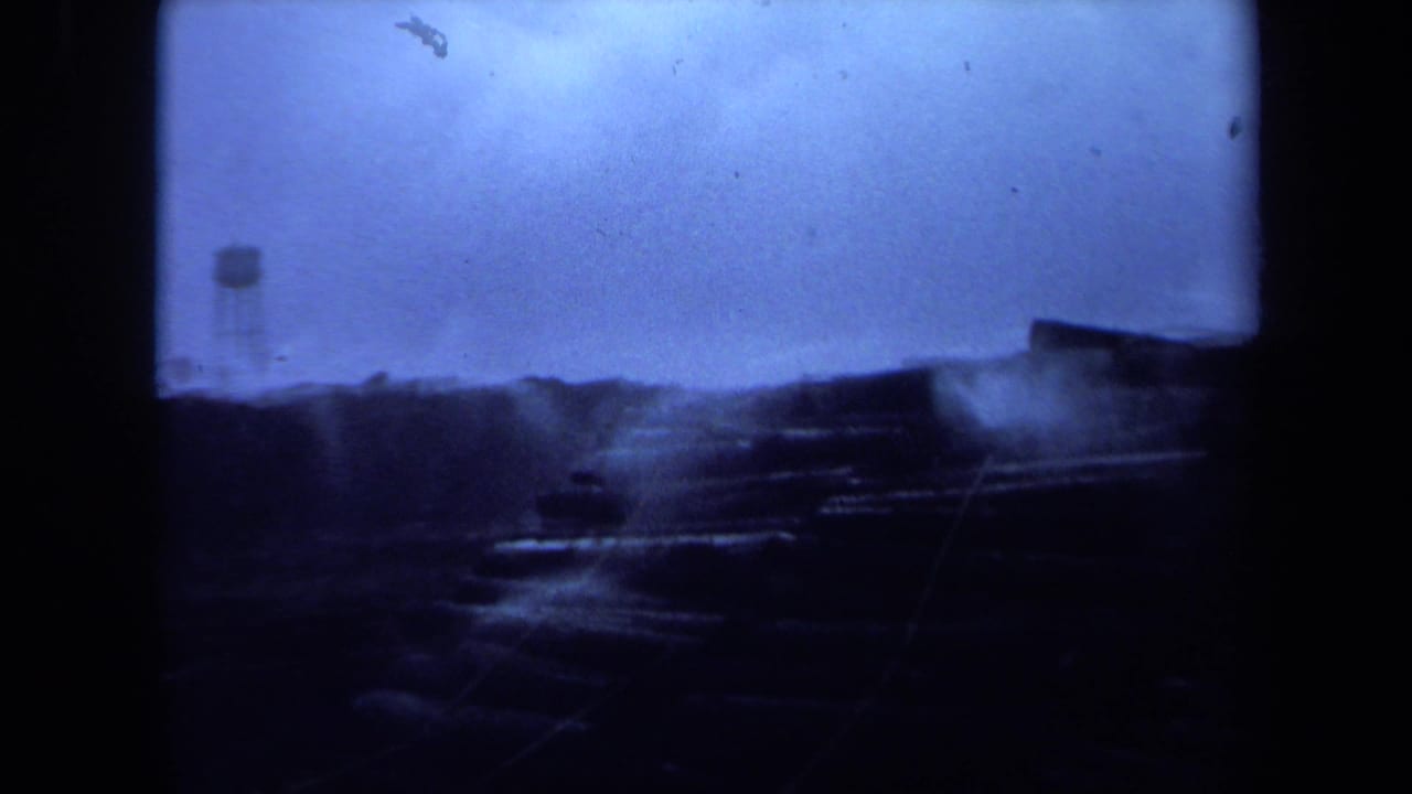 Dark waterfront view with a water tower, low structures, and blue overcast sky.
