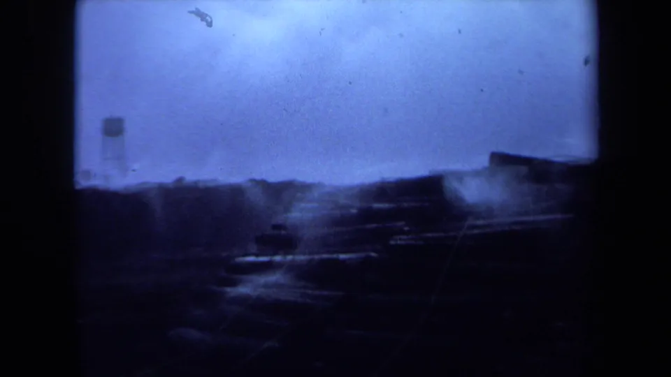 1971 Maine home-movie frame with a distant water tower beyond a dark shoreline and low waterfront structures in blue exposure.