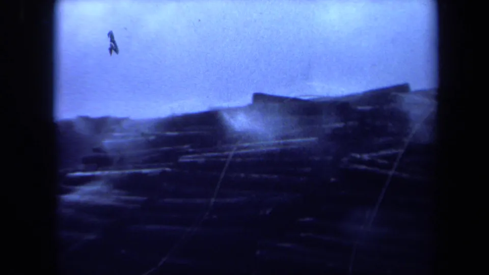 1971 Maine 8mm frame showing a damp, debris-strewn shoreline with low dark structures and visible scratches in the scan.