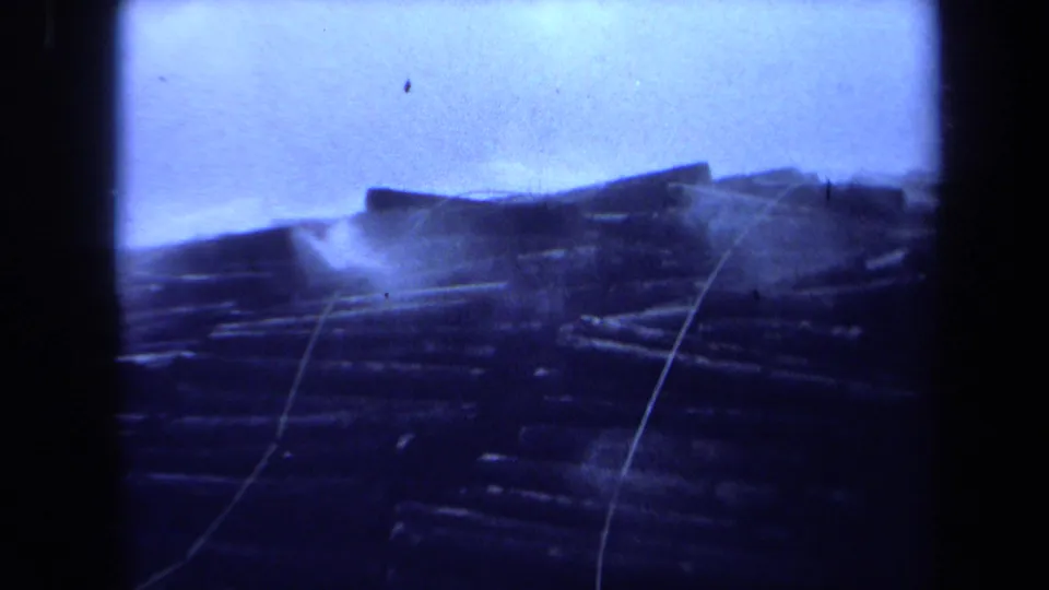 1971 Maine home-movie frame of a storm-dark shoreline with scattered debris, low buildings or structures, and prominent blue tint.