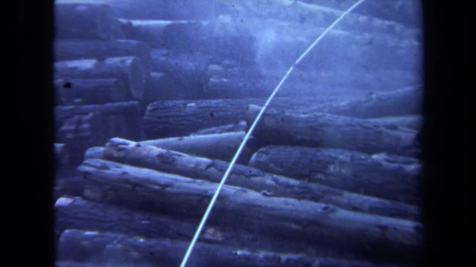 1971 Maine 8mm frame showing closer stacked logs, smoky haze, and a pale diagonal cable over the lumberyard scene.