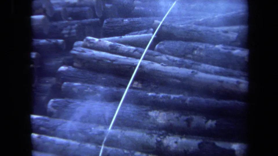 1971 Maine 8mm frame with layered lumberyard logs and a bright diagonal cable crossing the right half of the view.