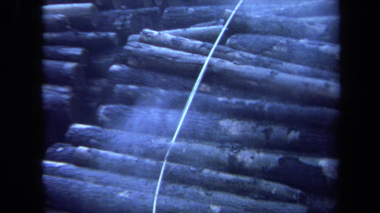 1971 Maine 8mm frame showing a pile of stacked logs outdoors with a pale line running diagonally across the scene.