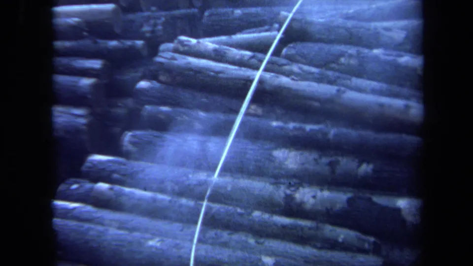 1971 Maine 8mm frame showing a pile of stacked logs outdoors with a pale line running diagonally across the scene.