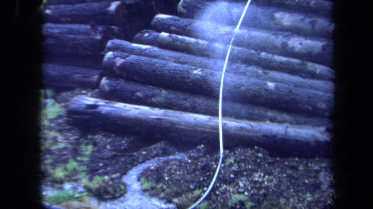 1971 Maine 8mm frame showing stacked logs near gravel, vegetation, and a narrow stream, with a pale cable running diagonally.