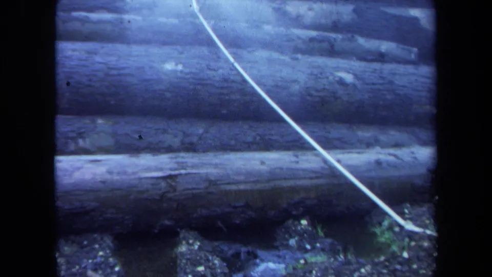1971 Maine 8mm frame with close stacked logs, gravelly ground, sparse vegetation, and a pale diagonal cable in front.